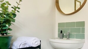Ein Badezimmer mit einem weißen Standwaschbecken, einem grünen Blumentopf, einem schwarzen Regal mit Handtüchern und einem Spiegel mit goldener Rahmung.