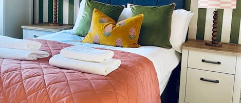Un lit bien fait, avec une couverture rose, deux oreillers et des serviettes pliées posées sur une table de chevet.