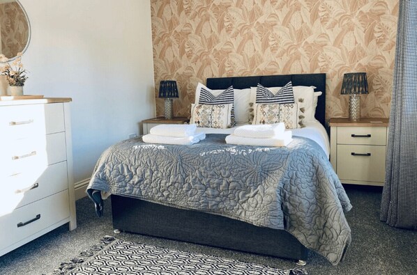 A neatly made bed with a grey floral duvet, two bedside tables with lamps, a mirror, and a patterned rug.