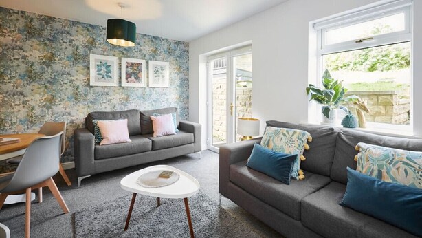 Een moderne woonkamer met een grijze bank, een ronde salontafel, en een eethoek met een houten tafel en stoelen. Er is een groot raam met uitzicht op groen.