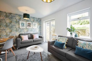 Een moderne woonkamer met een grijze bank, een ronde salontafel, en een eethoek met een houten tafel en stoelen. Er is een groot raam met uitzicht op groen.