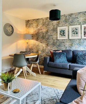 A modern living room with a grey sofa, a dining table with chairs, a clock, and framed botanical prints on the wall.