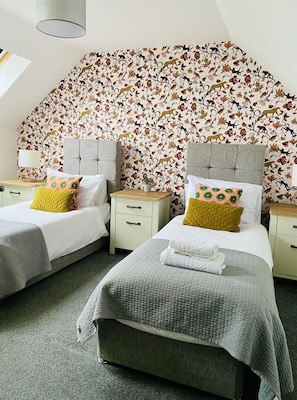 A hotel room with two beds, each with a grey headboard, white bedding, and a floral-patterned wallpaper. There are bedside tables with lamps and decorative pillows.