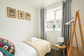 Une chambre à coucher avec un lit, une fenêtre avec des rideaux, un radiateur, un cintre en bois et des tableaux encadrés accrochés au mur.