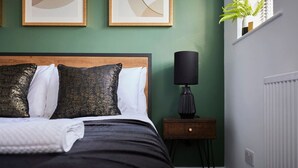Ein ordentlich gemachtes Bett mit dunklem Bettbezug und Kissen, ein kleiner Nachttisch mit Lampe sowie zwei gerahmte abstrakte Kunstwerke an der Wand.