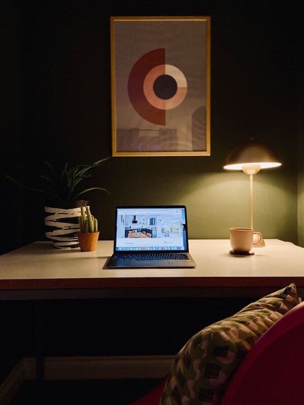 A laptop on a desk with a lamp, a potted plant, and a framed abstract artwork.
