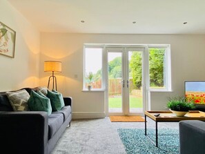 Relax in this inviting living space with a sofa, floor lamp, and TV, leading to a backyard with lush greenery.