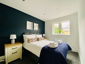Un lit bien fait, avec une couverture bleue, des oreillers blancs et un coussin à motifs en zigzag. Une table de chevet avec une lampe et une petite plante se trouve sur le rebord de la fenêtre.