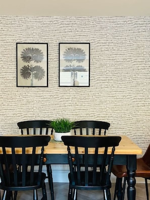 A dining area with a wooden table and black chairs, a potted plant on the table, and two framed pictures on the wall.
