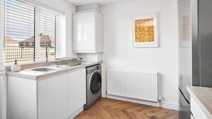 Une cuisine moderne avec des meubles blancs, un lave-linge encastré, un réfrigérateur en acier inoxydable et une fenêtre avec des stores.