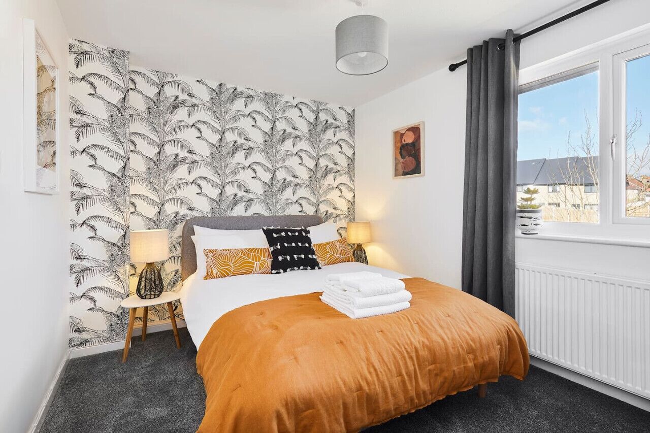 Habitación de hotel con una cama grande, dos lámparas de noche, una ventana con cortinas, un radiador y una pared con diseño de hojas.