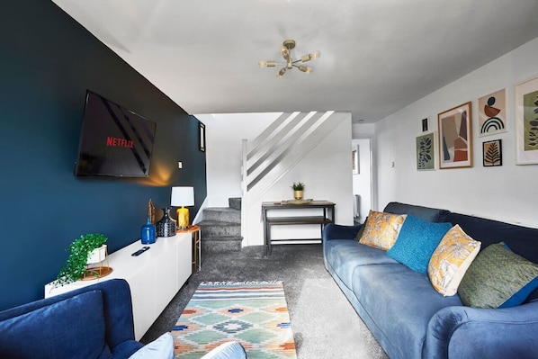 Una sala de estar moderna con un sofá azul, un televisor montado en la pared, una escalera y algunas obras de arte enmarcadas.