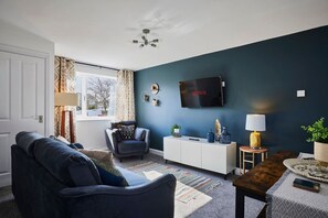 Un salon avec un mur bleu foncé, un téléviseur à écran plat, un canapé, un fauteuil, une table d’appoint et une lampe.