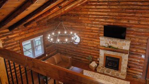 A log cabin interior with a stone fireplace, a television mounted above it, and a chandelier hanging from the ceiling.