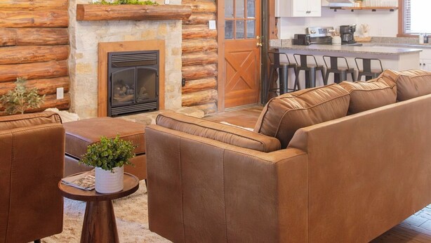 A log cabin interior with a fireplace, a sofa, a coffee table, and a kitchen area.