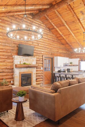 A log cabin interior with a fireplace, a sofa, a coffee table, and a kitchen area.