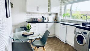 Cocina moderna con armarios blancos, una mesa de vidrio, dos sillas, una lavadora y un ventanal con vistas al verdor.