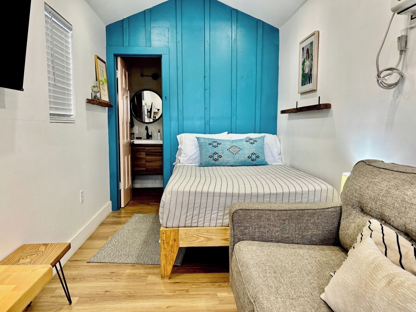 A bedroom with a bed, a wooden floor, a blue accent wall, a gray sofa, and a small table.