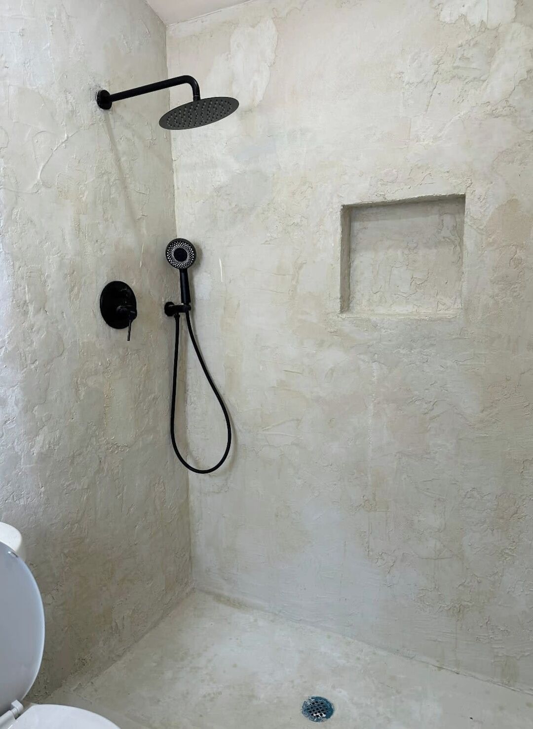 A shower with a handheld showerhead and a wall-mounted faucet in a room with a textured wall and a small square window.