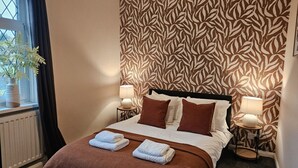 Un lit bien fait, avec une tête de lit à motifs marron et blanc, deux lampes de chevet et une fenêtre sur le rebord de laquelle est posée une plante.