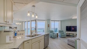 Una cocina moderna con una isla grande, lavavajillas y un fregadero. Hay un comedor con una silla y un televisor. La habitación tiene ventanas amplias y vista al océano.