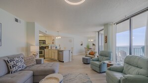 Amplia sala de estar con sofá, sillones y una mesa de comedor. Se ve una cocina al fondo, y un balcón con vistas.