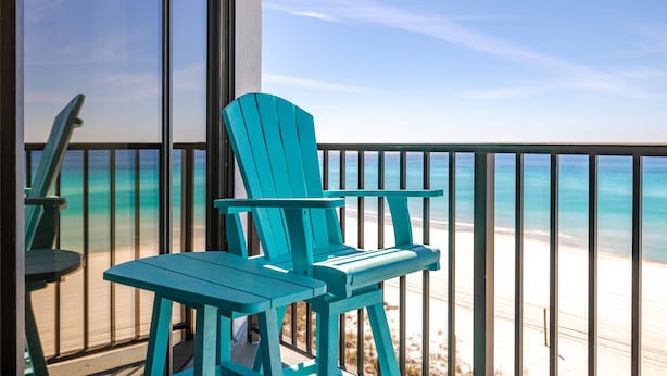 Un balcón con sillas Adirondack turquesa y una mesa, con vista a una playa de aguas azules cristalinas y orilla arenosa.