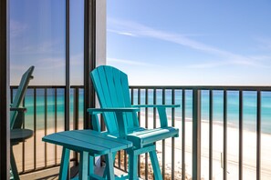 A balcony with turquoise Adirondack chairs and a table, overlooking a beach with clear blue water and sandy shore. - 503-E Free Beach Service Lounge Chairs + Umbrella (Panama City Beach)