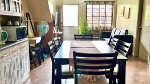 A dining area with a long table, chairs, a fan, and kitchen appliances. - Dovilim Apartments (Tacloban)