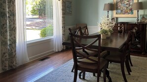 A dining room with a wooden table and chairs, a chandelier, and a view of the outdoors through a window with floral curtains. - Charming spacious home nestled on a private lot minutes away from the Masters (Augusta)