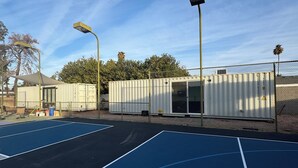 Ein Basketballplatz im Freien mit blau-grauen Markierungen, umgeben von einem Maschendrahtzaun und Metallgebäuden.
