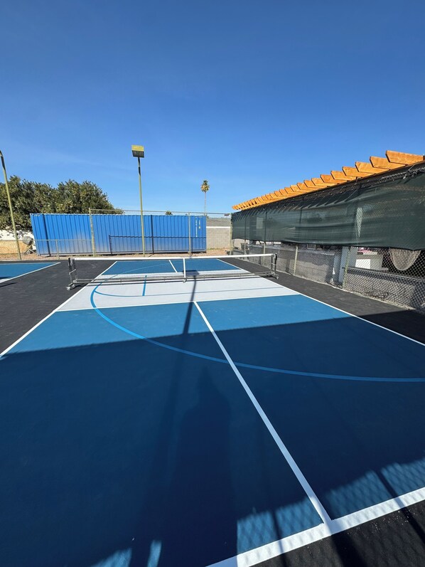 An outdoor tennis court with blue and black surfaces, surrounded by a chain-link fence and a blue metal fence. - Charming 1-bedroom casita in peaceful Mesa with pickleball sports court. (Mesa)