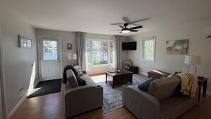 A modern living room with a sofa, a coffee table, and a television. There are two windows with curtains, a ceiling fan, and a door. - Riverside Landing Cottage (Alberton)