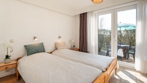 Ein Schlafzimmer mit zwei Betten, einem hölzernen Kopfteil, einem Nachttisch mit Lampe, einer Zimmerpflanze und Blick ins Freie durch eine Schiebetür.