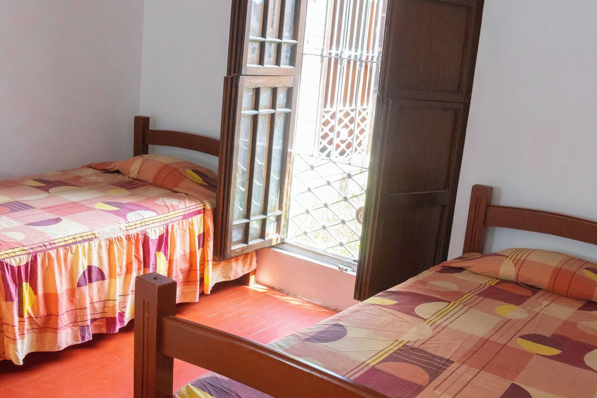 A room with two wooden beds with colorful bedding, a wooden door, and a window with metal bars.