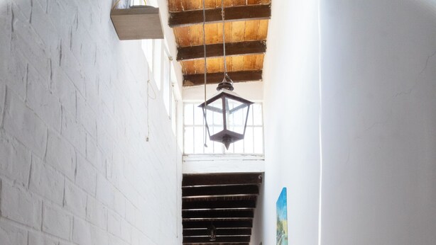 A narrow corridor with white brick walls and a red brick floor, leading to a doorway with a view of greenery outside. - Hawka (Ica)