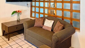 Ein gemütliches Zimmer mit Holzbalkendecke, einem an der Wand montierten Fernseher, einem Fenster mit Holzrahmen, einer Couch mit Kissen und einem kleinen Tisch mit einer Blumenvase.