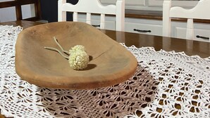 Ein Esstisch mit einer hölzernen Schüssel als Mittelstück, einer Spitzen Tischläufer und einem weißen Schrank mit Glastüren im Hintergrund.