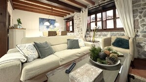 Ein gemütliches Wohnzimmer mit einer Couch, einem Couchtisch und einer Steinwand. Ein Fenster mit Vorhängen, ein Gemälde an der Wand und eine Pendelleuchte sind zu sehen.