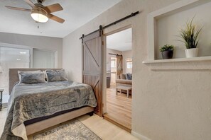 A bedroom with a bed, a ceiling fan, a sliding barn door, a small table, and a shelf with plants.