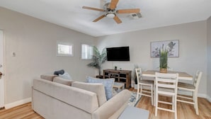 Een woonkamer met een beige bank, een houten eettafel met stoelen, een plafondventilator en een flatscreen televisie die aan de muur is bevestigd.