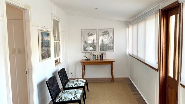 A hallway with wooden flooring, a sofa, two chairs with patterned cushions, a small table with books, and framed pictures on the wall. - The Merchant's Nook (Armidale)