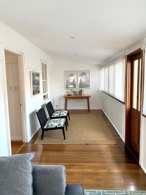 A hallway with wooden flooring, a sofa, two chairs with patterned cushions, a small table with books, and framed pictures on the wall. - The Merchant's Nook (Armidale)