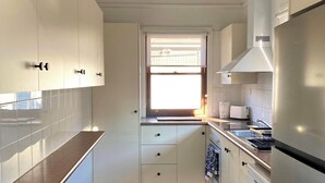 A compact kitchen with white cabinets, a stainless steel refrigerator, and a window with blinds. - The Merchant's Nook (Armidale)