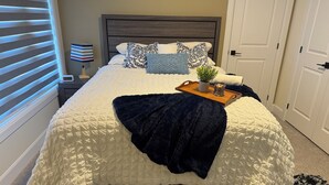 Uma cama bem arrumada com um cobertor escuro, uma mesa de cabeceira com um relógio e uma pequena planta em um suporte.