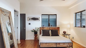 Un dormitorio con una cama de madera, ventilador de techo, ventana con persianas, un cuadro enmarco con un pez y un espejo.