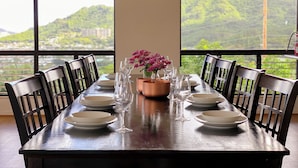 Una mesa de comedor para seis personas con sillas, un florero con flores rosadas y vistas a colinas verdes a través de grandes ventanales.