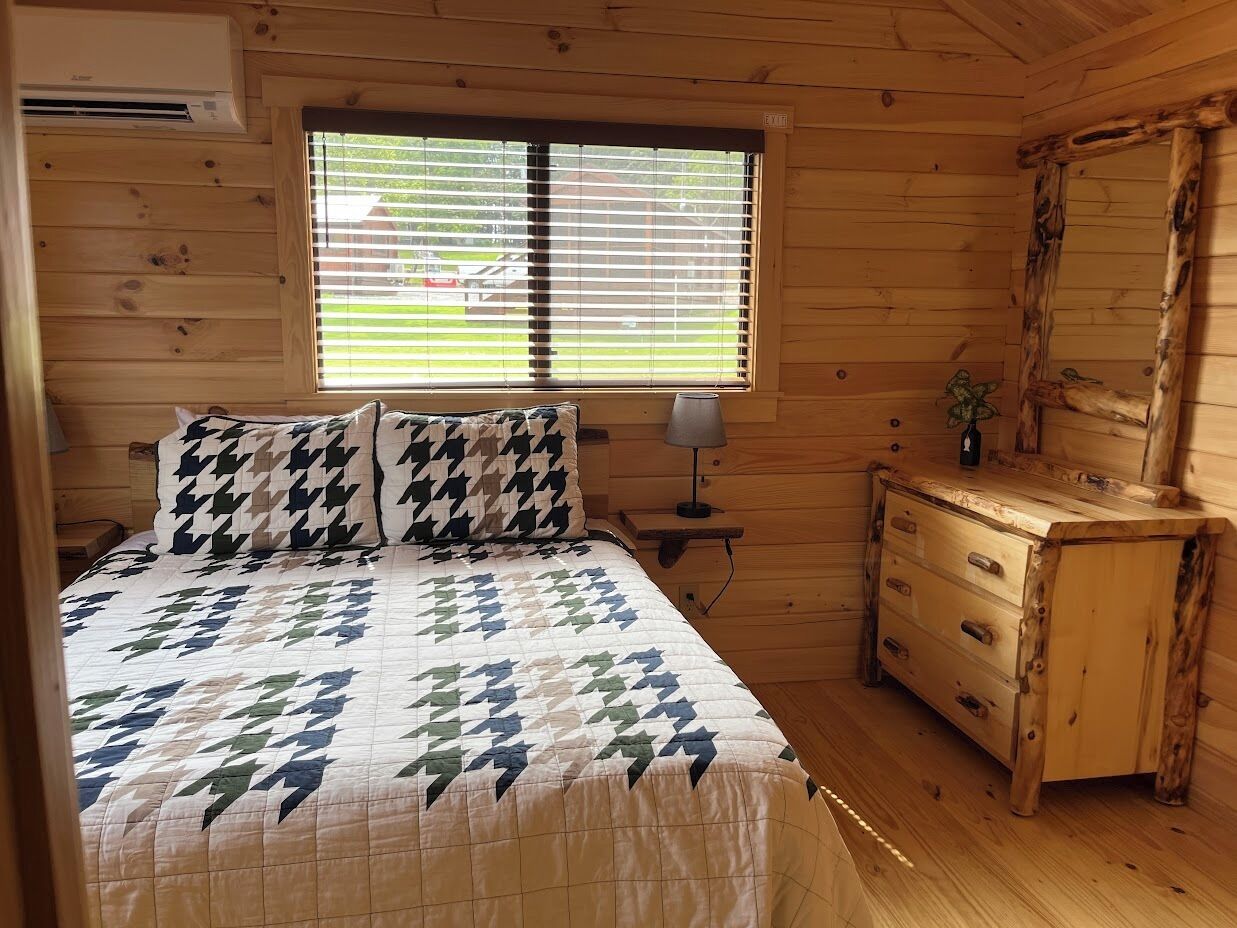 Quarto de cabana de madeira com cama, mesa de cabeceira, cômoda e janela com persianas.