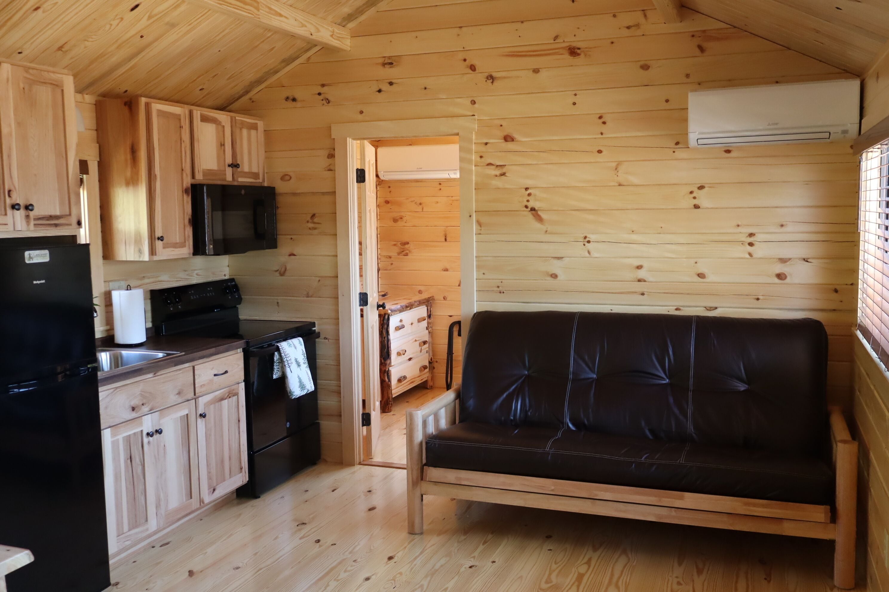 A wooden cabin interior with a kitchenette, a black leather sofa, and a wooden dining table.