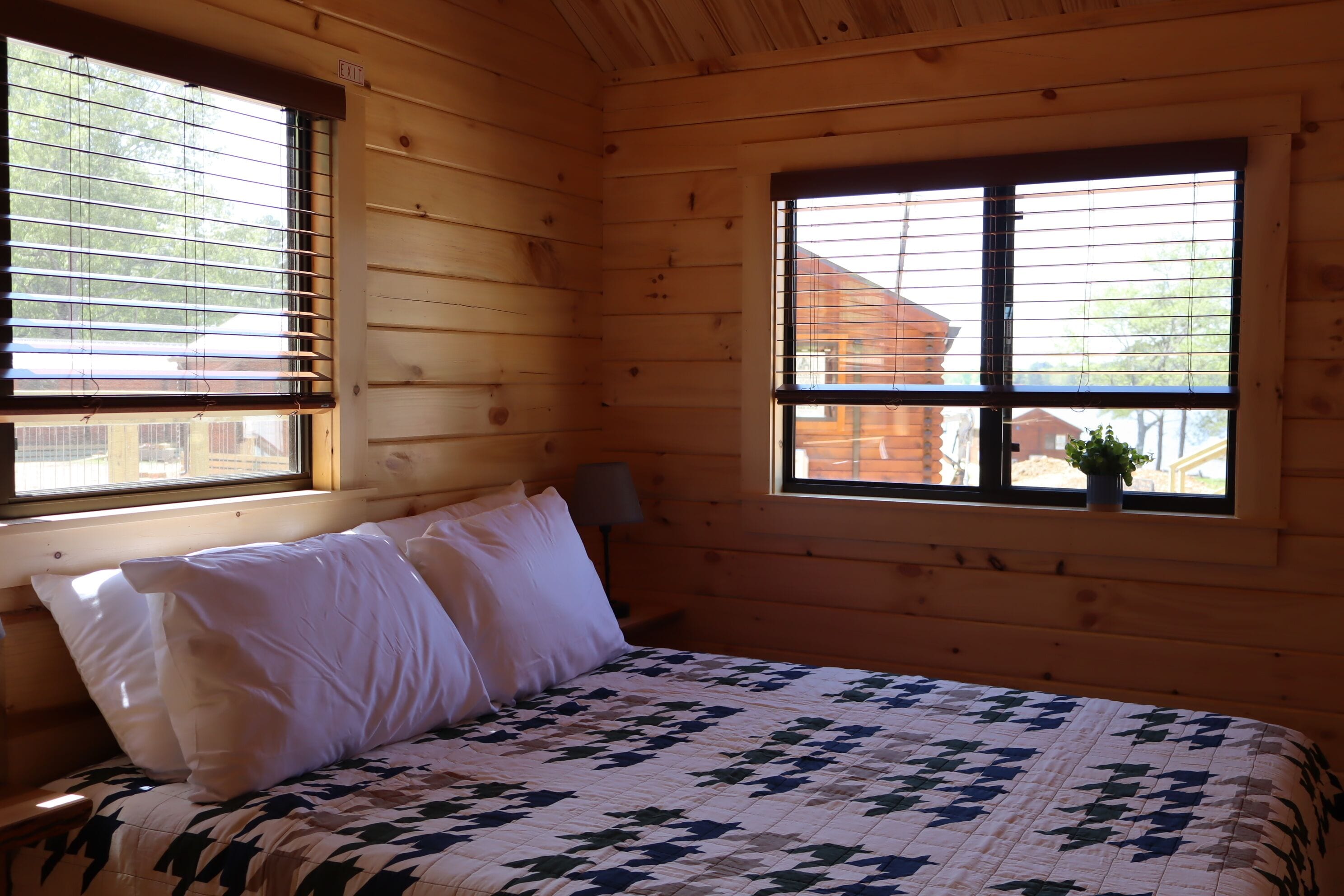 Une chambre dans une cabane en bois, avec un lit, deux fenêtres et une lampe.